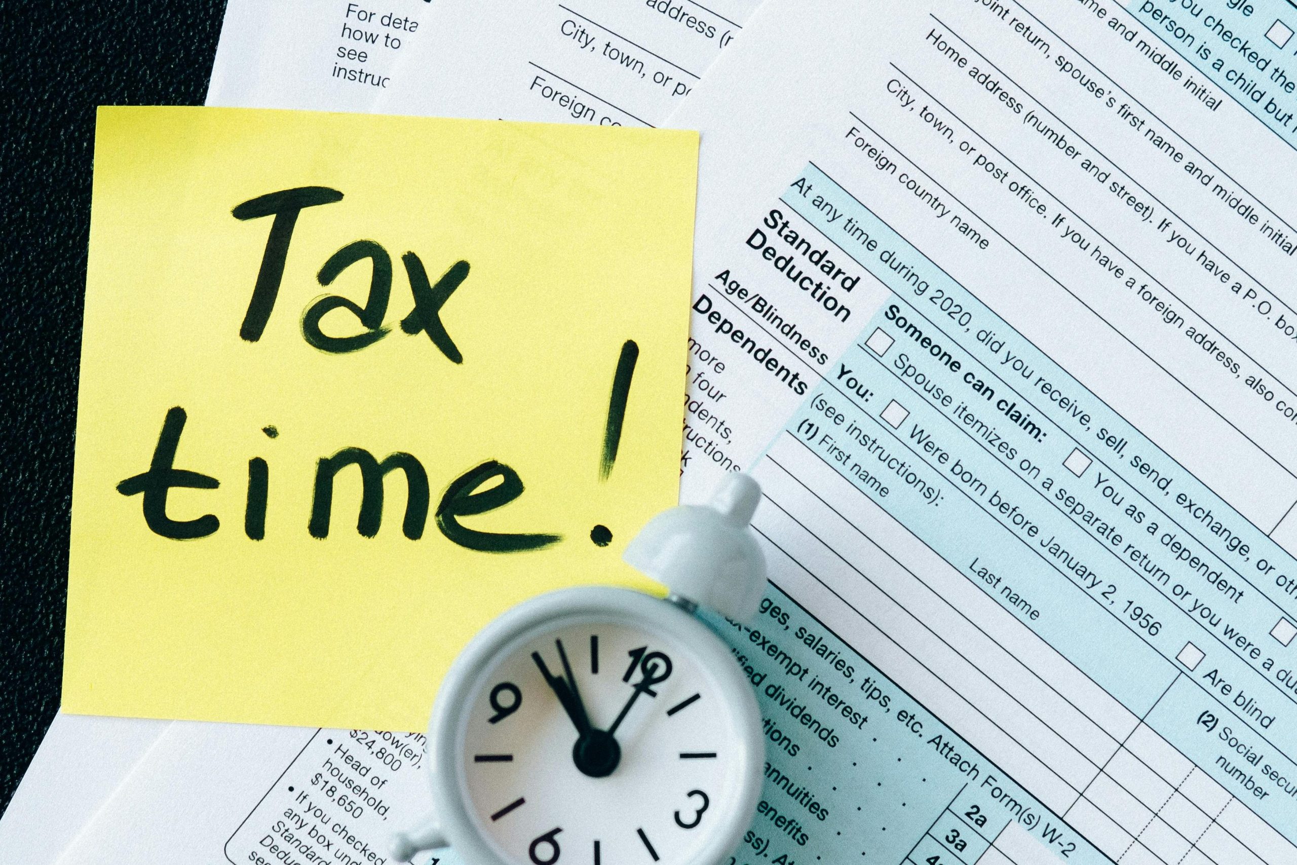 A stack of tax forms with a clock and yellow sticky note saying 'Tax time!' indicating urgency.